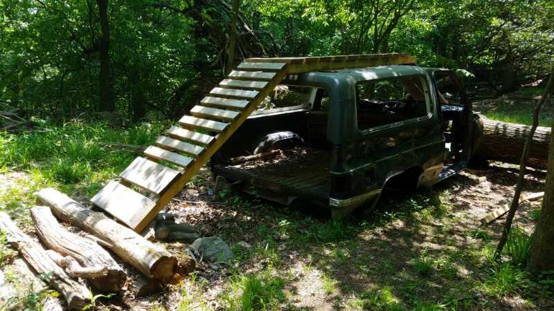 An abandoned vehicle partially covered by trees in a forested area, with a wooden ramp leading up to it and logs scattered nearby on the ground. Sprain Ridge mountain bike trail.