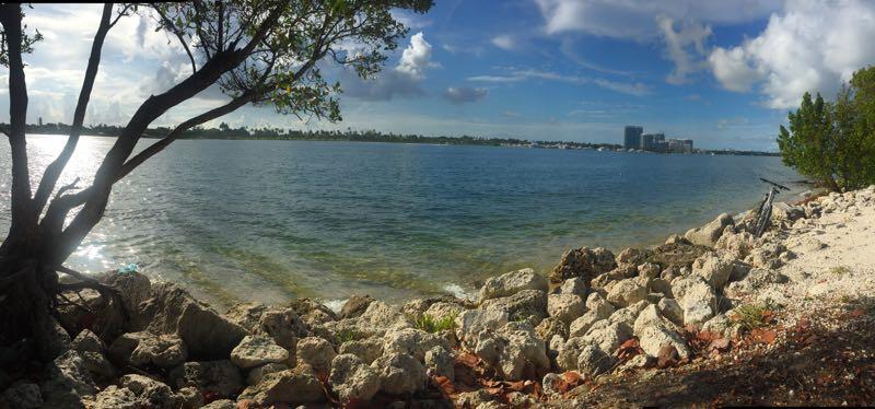 A panoramic view of a calm waterfront scene, featuring clear blue water and a rocky shore lined with small stones. On the left, a tree casts a shadow, and on the right, lush greenery borders the beach. In the background, city buildings are visible against a partly cloudy sky, creating a serene and picturesque landscape. Oleta River State Park mountain bike trail.