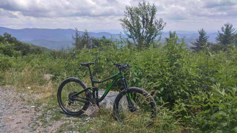 A mountain bike resting on a rocky outcrop surrounded by tall grass and shrubs, with a scenic view of rolling hills and mountains in the background under a partly cloudy sky. Bear Creek mountain bike trail.