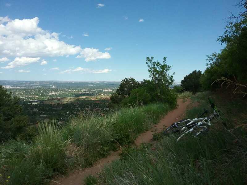 A scenic view from a hillside trail, featuring a dirt path surrounded by green grass and shrubs. In the foreground, a white and green bicycle is leaned against the edge, while the background showcases a broad landscape with distant mountains and a blue sky dotted with white clouds. Paul Intemann Memorial Trail mountain bike trail.