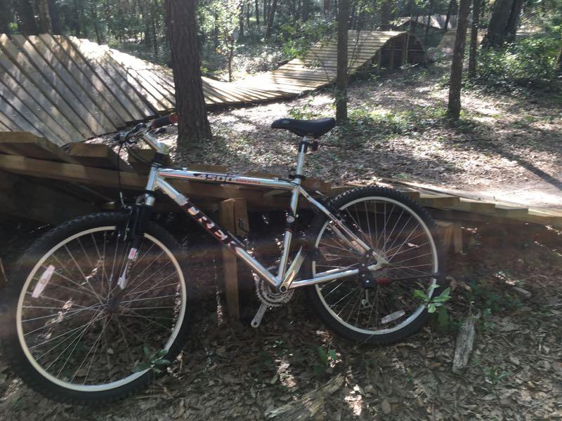A mountain bike leaning against a wooden structure, with a dirt path and trees in the background, indicating a biking trail. Sunlight filters through the trees, casting dappled shadows on the ground. Santos mountain bike trail.
