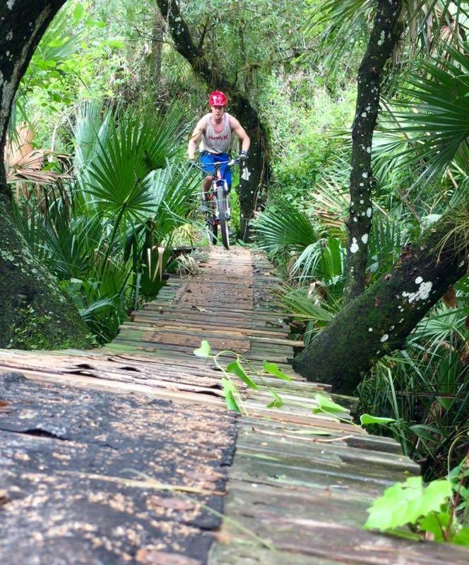 A person riding a mountain bike on a narrow wooden bridge surrounded by lush greenery and tropical plants. The rider is wearing a red helmet and a tank top, navigating through a dense forest area. North Port Mountain Bike Trails mountain bike trail.