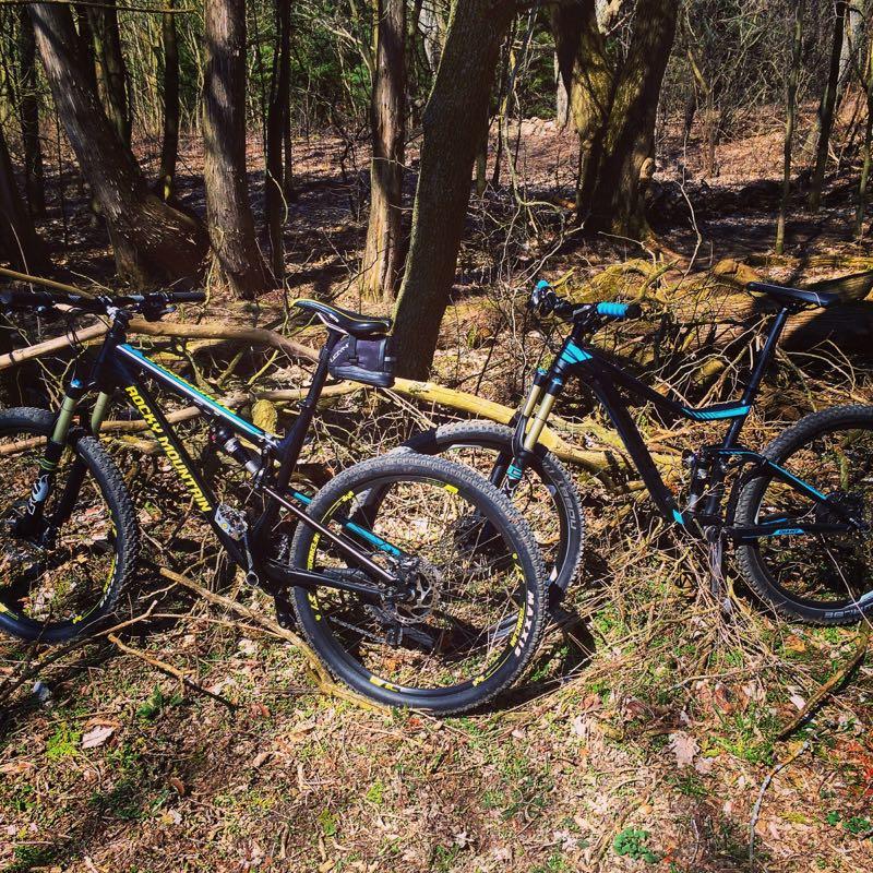 Two mountain bikes are parked beside a fallen tree in a wooded area with sparse foliage and sunlight filtering through the trees. The bikes are black with colorful accents and are resting on the ground covered in leaves and grass. The Pines mountain bike trail.