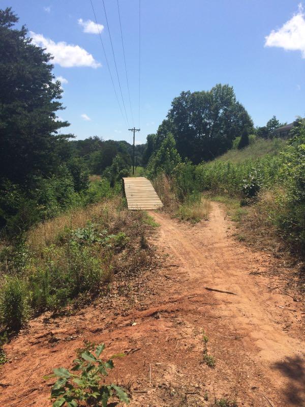 A dirt path winding through a lush, green area, leading to a wooden bridge over a small ditch. Utility poles are visible in the background under a blue sky with a few clouds. Signal Hill mountain bike trail.