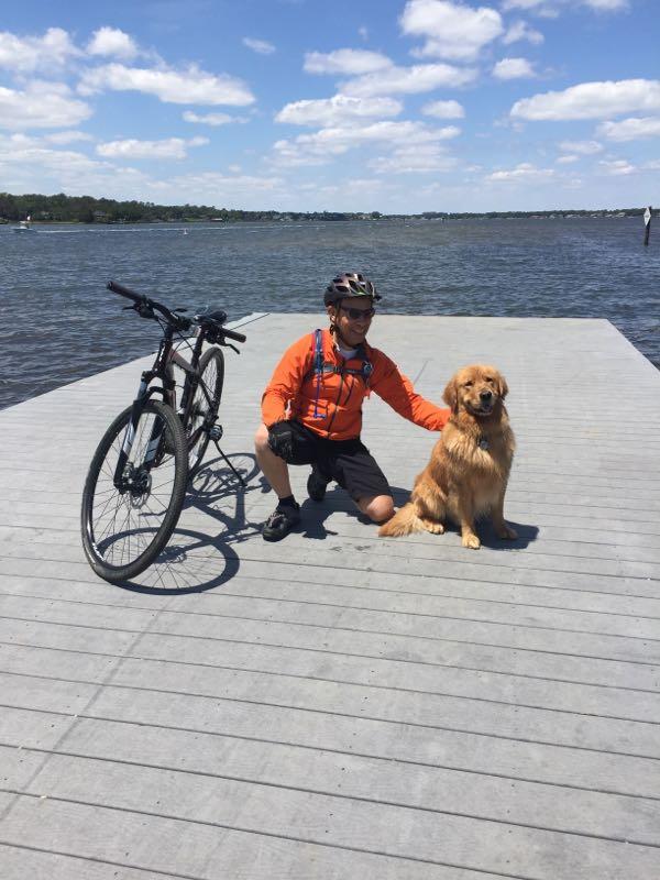 A person in an orange cycling jacket kneels next to a golden retriever on a wooden dock by a lake. A bicycle is parked nearby, and the sky is partly cloudy. First Landing State Park mountain bike trail.