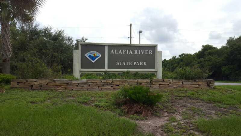 Sign for Alafia River State Park with a landscaped area featuring grass and shrubs. The sign displays the park's name and a blue emblem. Alafia River State Park mountain bike trail.