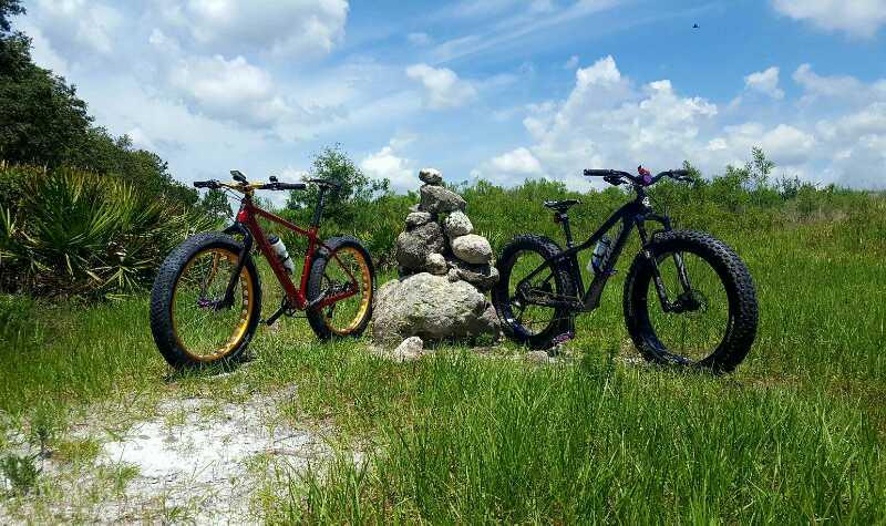 Two fat tire bicycles, one red and the other black, stand beside a rock cairn in a grassy field under a bright blue sky with clouds. The background features lush greenery, creating a scenic outdoor environment. Balm Boyette Scrub Preserve mountain bike trail.