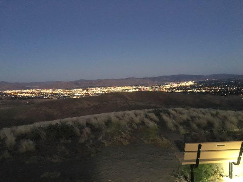 A scenic view of a city illuminated at night, taken from a hillside. In the foreground, there is a bench positioned to overlook the lights of the city below, with hills and distant mountains framing the background. The sky is darkening, showcasing the transition from dusk to night. Keystone Canyon mountain bike trail.