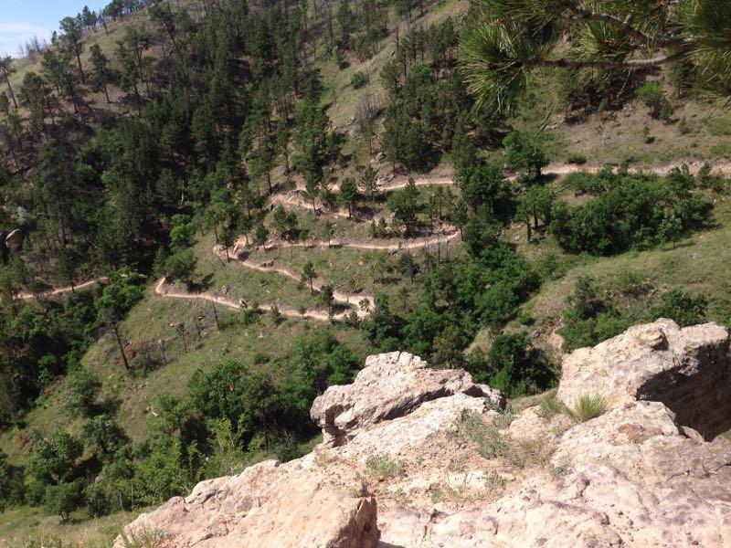 Alt text: A winding dirt trail navigates through a lush green landscape with patches of trees and rocky outcrops, viewed from a high vantage point. HLMP mountain bike trail.