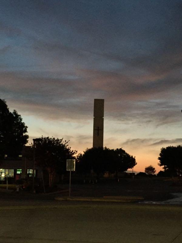 A silhouette of a church tower with a cross at sunset, surrounded by trees and a quiet parking area. The sky displays shades of blue, purple, and orange as the day transitions to night. Northshore Trail mountain bike trail.