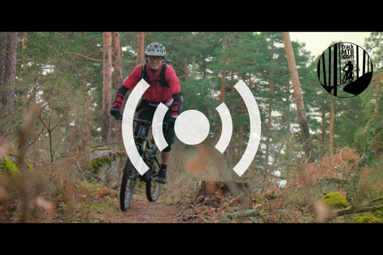 A cyclist in a red jersey and helmet rides a mountain bike on a wooded trail surrounded by trees and foliage. An overlay symbol indicating sound or connectivity is present in the center, along with a logo in the top right corner for trackMTB.com.
