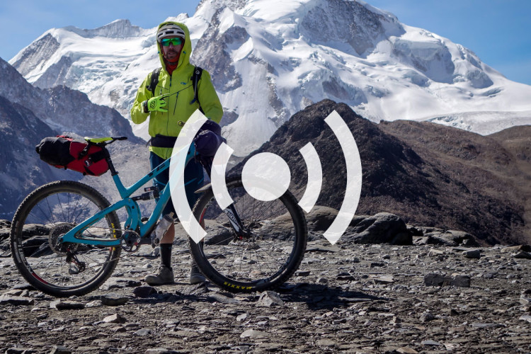 A mountain biker stands beside a blue bicycle on rocky terrain, wearing a bright yellow jacket and helmet, with snow-capped mountains in the background under a clear blue sky.