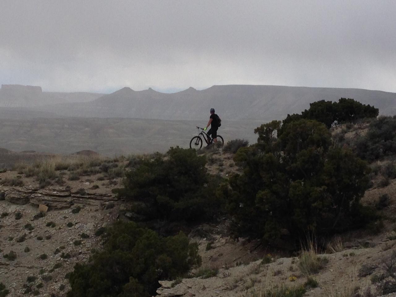 A mountain biker pauses on a rocky trail overlooking a vast, rugged landscape with rolling hills and distant mountains under a cloudy sky. The scene features sparse vegetation, including shrubbery and grasses, set against a backdrop of gray clouds. Wilkins Peak Trails mountain bike trail.
