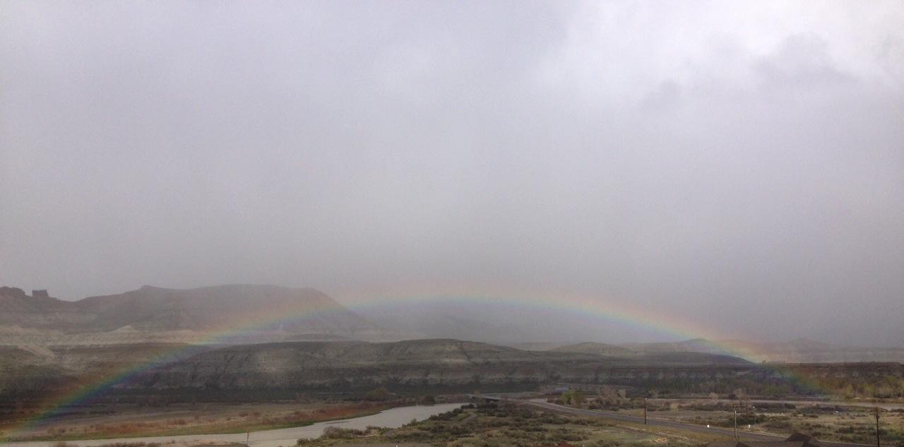 A rainbow arcs over a mountainous landscape under a cloudy sky, with a river and a winding road visible below. The scene captures a blend of muted grey tones and the vibrant colors of the rainbow. Wilkins Peak Trails mountain bike trail.