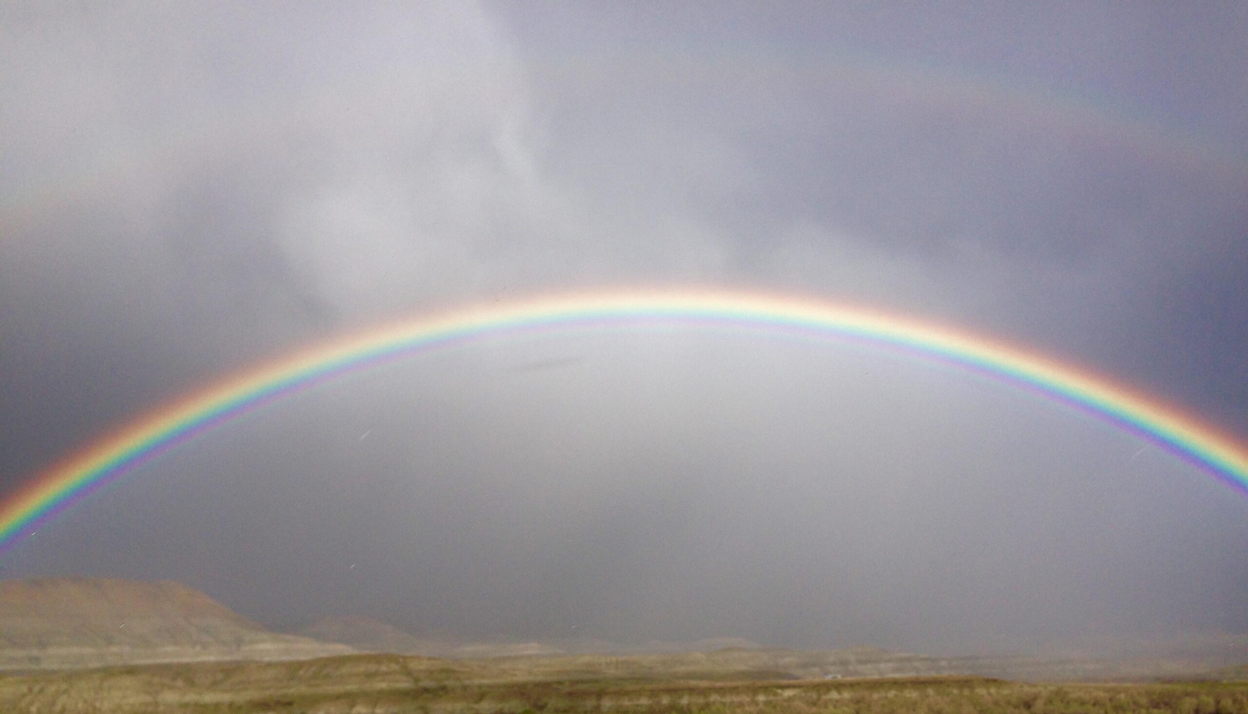 A vibrant rainbow arcs across a cloudy sky, with shades of red, orange, yellow, green, blue, and violet visible. The landscape below features rolling hills and a muted, earthy color palette, enhancing the brightness of the rainbow against the gray backdrop. Wilkins Peak Trails mountain bike trail.