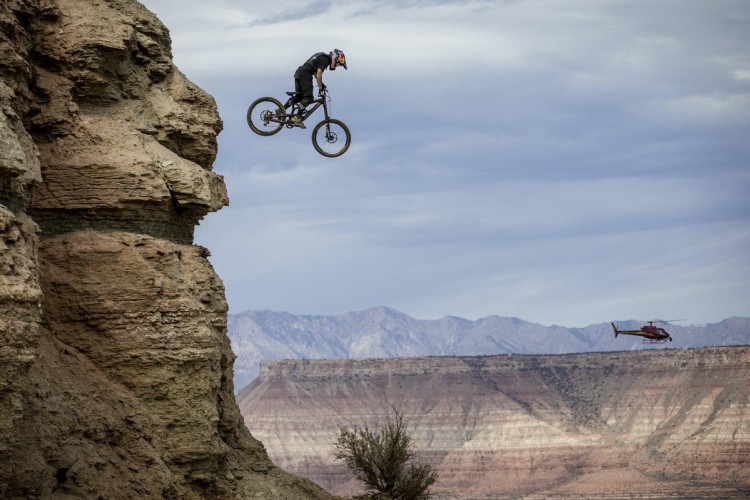 A mountain biker performs a jump off a rocky cliff, with a helicopter flying in the background against a dramatic landscape of mountains and a cloudy sky.