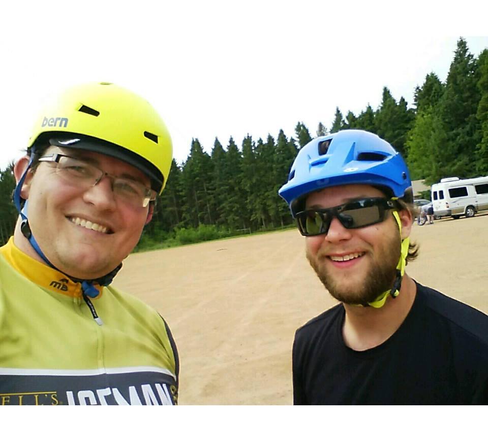 Two smiling men pose for a selfie outdoors, both wearing helmets. One man has a bright yellow helmet, glasses, and a cycling jersey. The other has a blue helmet, sunglasses, and a dark shirt. They are in a grassy area with trees in the background. Nine Mile mountain bike trail.