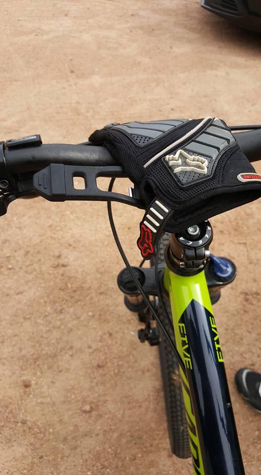 A close-up view of a mountain bike handlebar featuring a black and gray glove resting on the grip. The bike frame is predominantly green with the word "FIVE" printed multiple times. The background shows a dirt surface and part of a vehicle. Nine Mile mountain bike trail.