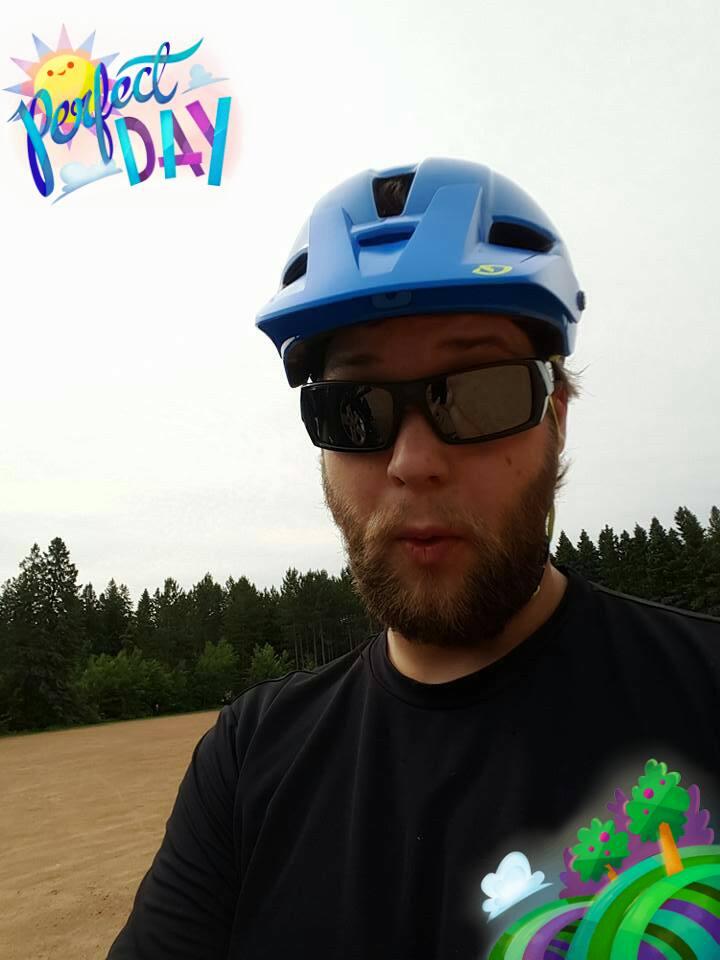 A person wearing a blue bike helmet and black sunglasses stands outdoors, with trees in the background. They have a surprised expression and are dressed in a black shirt. The image includes playful graphics that read "Perfect Day" along with colorful elements like trees and clouds. Nine Mile mountain bike trail.