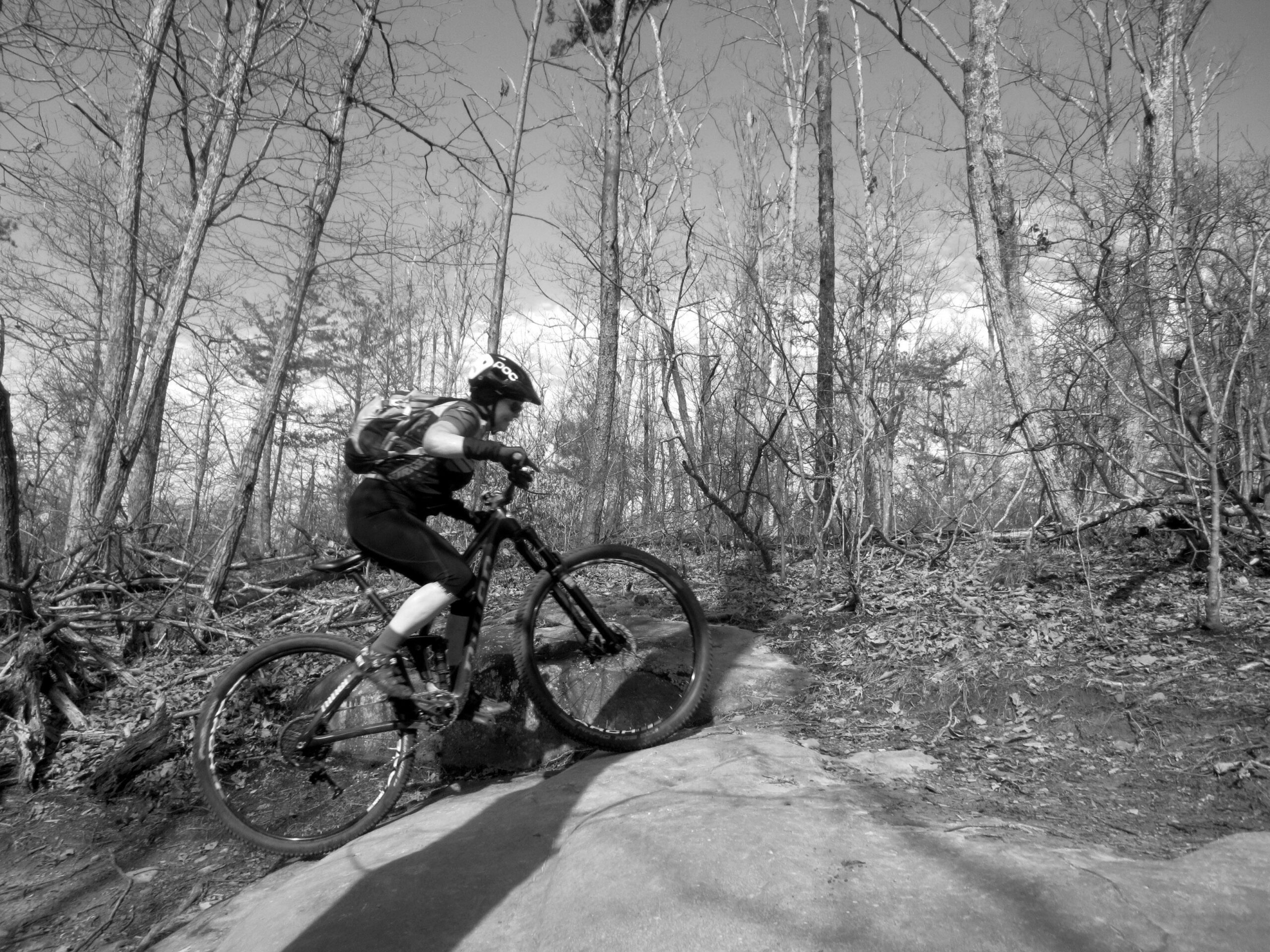 Niner Jet 9 RDO: A cyclist navigating a rocky trail in a wooded area, captured in black and white. The rider is mid-motion, climbing over a large rock, surrounded by bare trees and dry leaves.