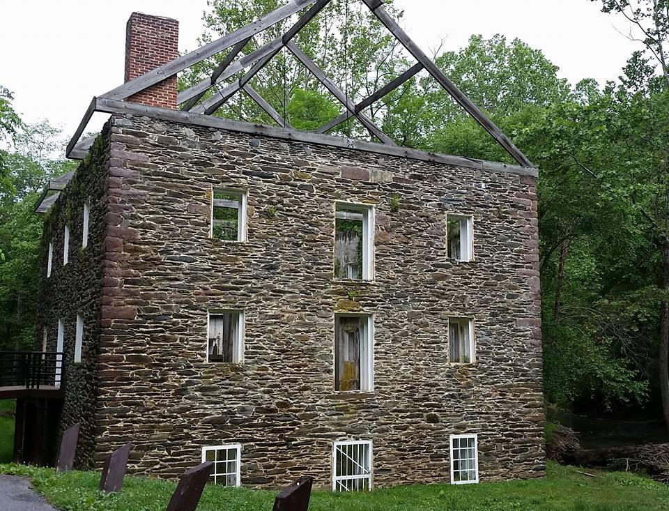 An old stone building with a partially collapsed roof surrounded by green foliage. The structure features several empty window frames and a rustic chimney. In the foreground, there is a grassy area with steps leading to the building, and a small stream visible to the side. Schaeffer Farms mountain bike trail.