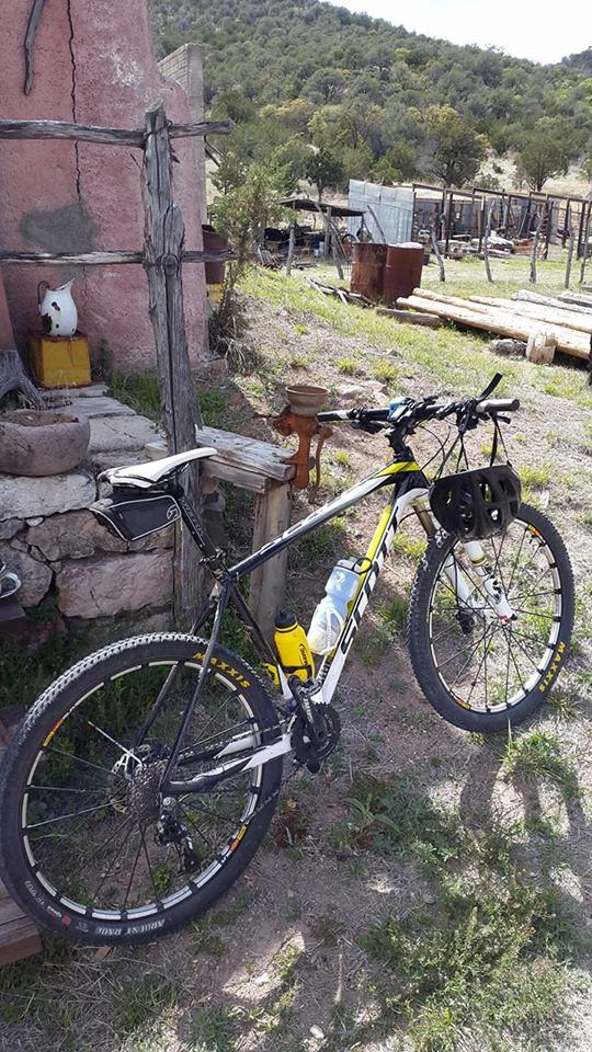 Scott Scale 920: A black and yellow mountain bike is leaning against a rustic wooden fence, with a small stone structure visible in the background. The area features dry grass and scattered rocks, surrounded by rolling hills and trees. A water bottle and other gear are attached to the bike.