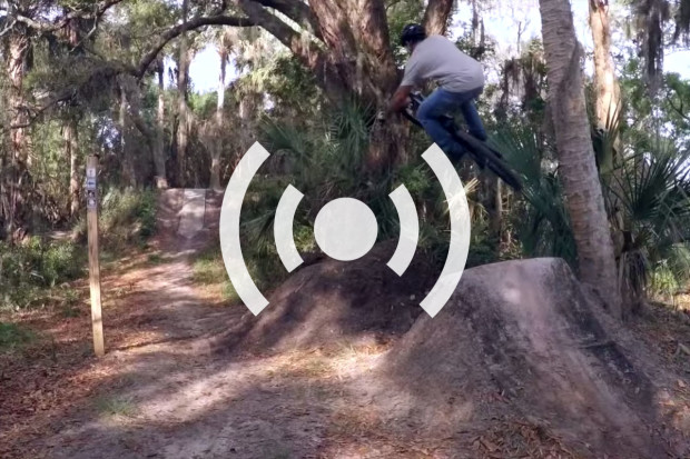 A mountain biker performing a jump over a dirt ramp in a wooded area, surrounded by trees and greenery, with a trail visible in the background. An audio symbol is superimposed on the image.