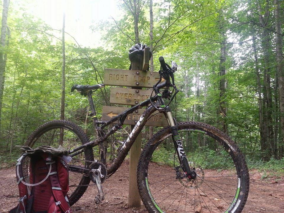 Mountain bike leaning against a wooden trail sign in a lush forest. The sign indicates different trail loops, and a helmet is placed on top of the sign. A backpack is visible on the ground near the bike, with dirt and mud on the bike's tires, suggesting recent use on the trails. Tsali Recreation Area mountain bike trail.