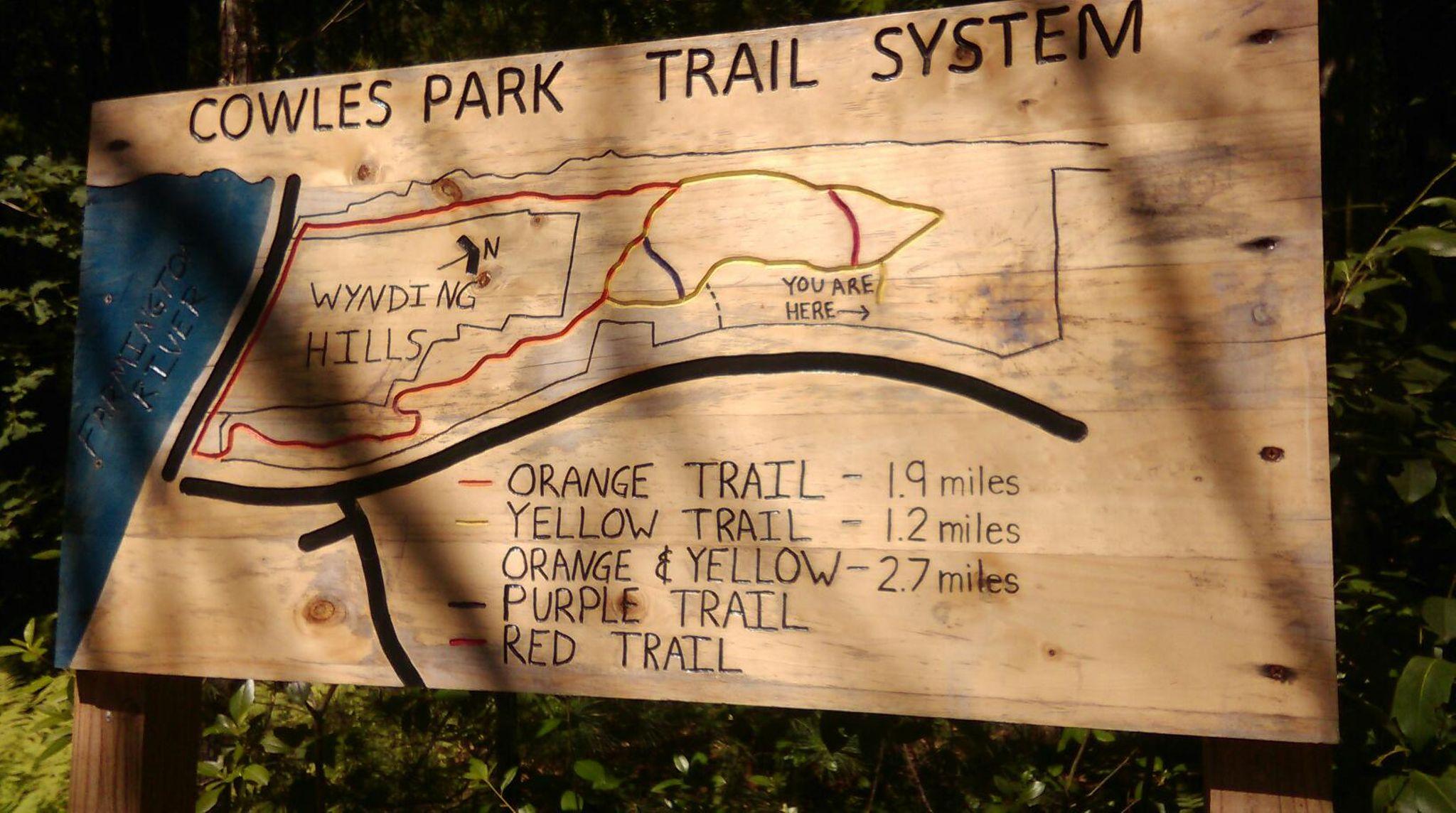 A wooden trail map sign for Cowles Park Trail System, featuring a map with labeled trails including Orange, Yellow, Purple, and Red, along with distances for each trail. The sign indicates "You Are Here" and includes a directional arrow pointing north. Surrounding greenery adds a natural backdrop. Cowles Park mountain bike trail.