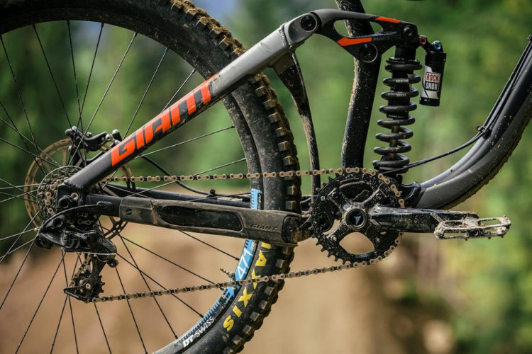 A close-up of a mountain bike's rear section showcasing the drivetrain, suspension, and frame. The frame prominently displays the brand name "Giant" in bold lettering. The components include a black chainring, rear derailleur, and a coil shock absorber from RockShox, all mounted on a dirt-covered bicycle with a knobby tire. The background features a blurred natural landscape, suggesting an outdoor biking environment.