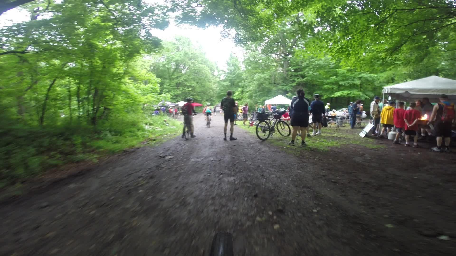 A dirt path surrounded by lush greenery, with several people engaging in activities related to a biking event. In the background, tents are set up, and groups gather, including cyclists and spectators. The scene conveys a lively outdoor atmosphere during a bike race or festival. Stewart State Forest mountain bike trail.