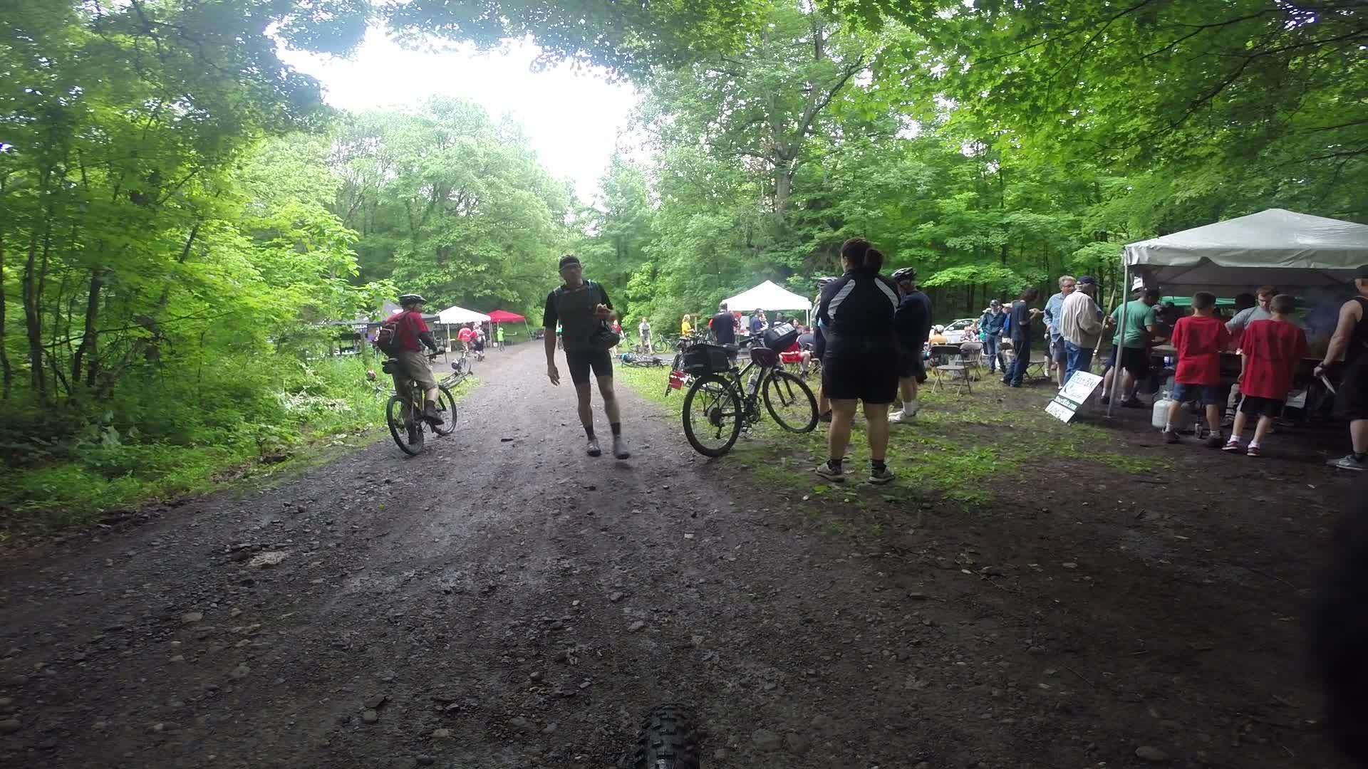 A dirt path lined with greenery where several people are gathered near tents. Some individuals are walking or riding bicycles, while others are socializing and enjoying the outdoor event. Stewart State Forest mountain bike trail.