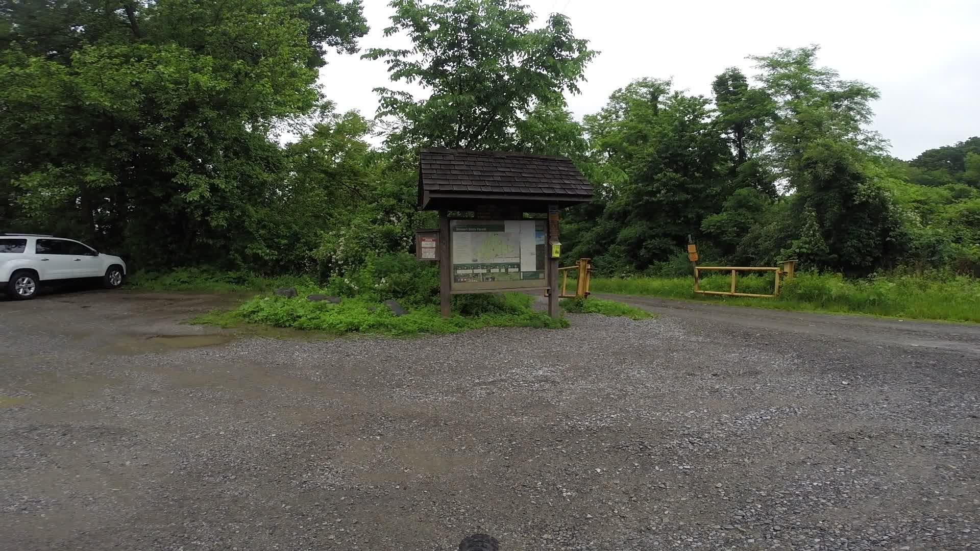 A gravel parking area with a white SUV on the left, featuring a wooden information kiosk displaying a map and signs. Lush greenery surrounds the area, indicating a natural setting, with a yellow gate visible to the right. The scene is slightly overcast, adding a serene atmosphere. Stewart State Forest mountain bike trail.