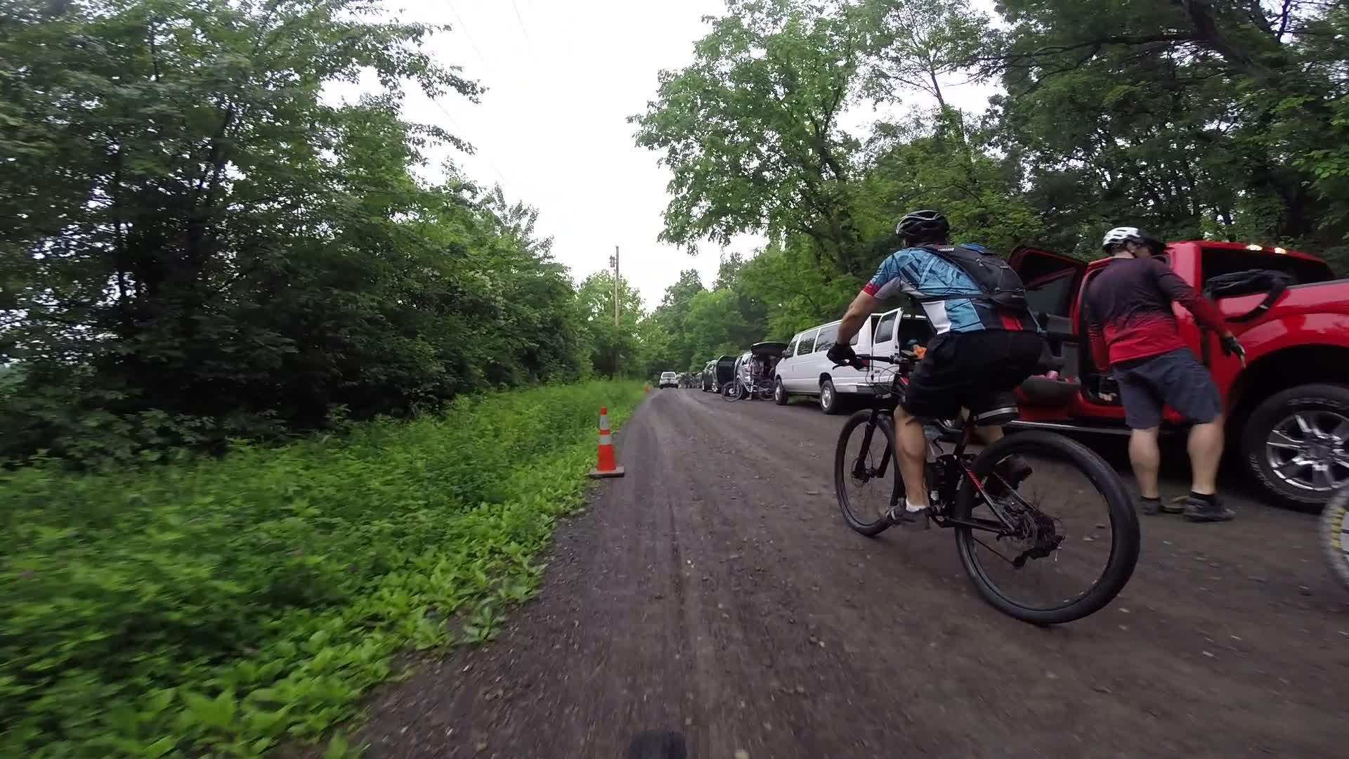 A cyclist rides on a gravel path alongside parked vehicles, with dense greenery on both sides. There are traffic cones marking the pathway, and another person is seen interacting with the rear of a red truck. The scene is set in a natural, outdoor environment on a cloudy day. Stewart State Forest mountain bike trail.