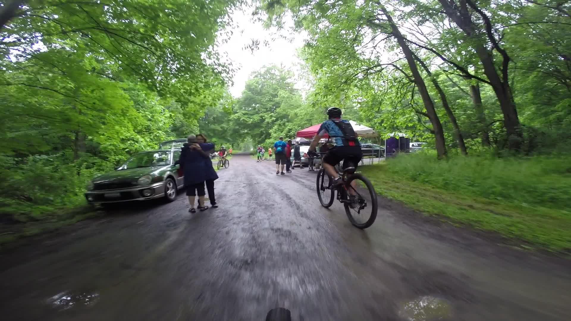 A cyclist riding on a gravel path surrounded by lush green trees, with a group of people gathered nearby, including two individuals embracing. There are cars parked along the side of the path, and a canopy tent set up in the background, indicating a social or community event. Stewart State Forest mountain bike trail.