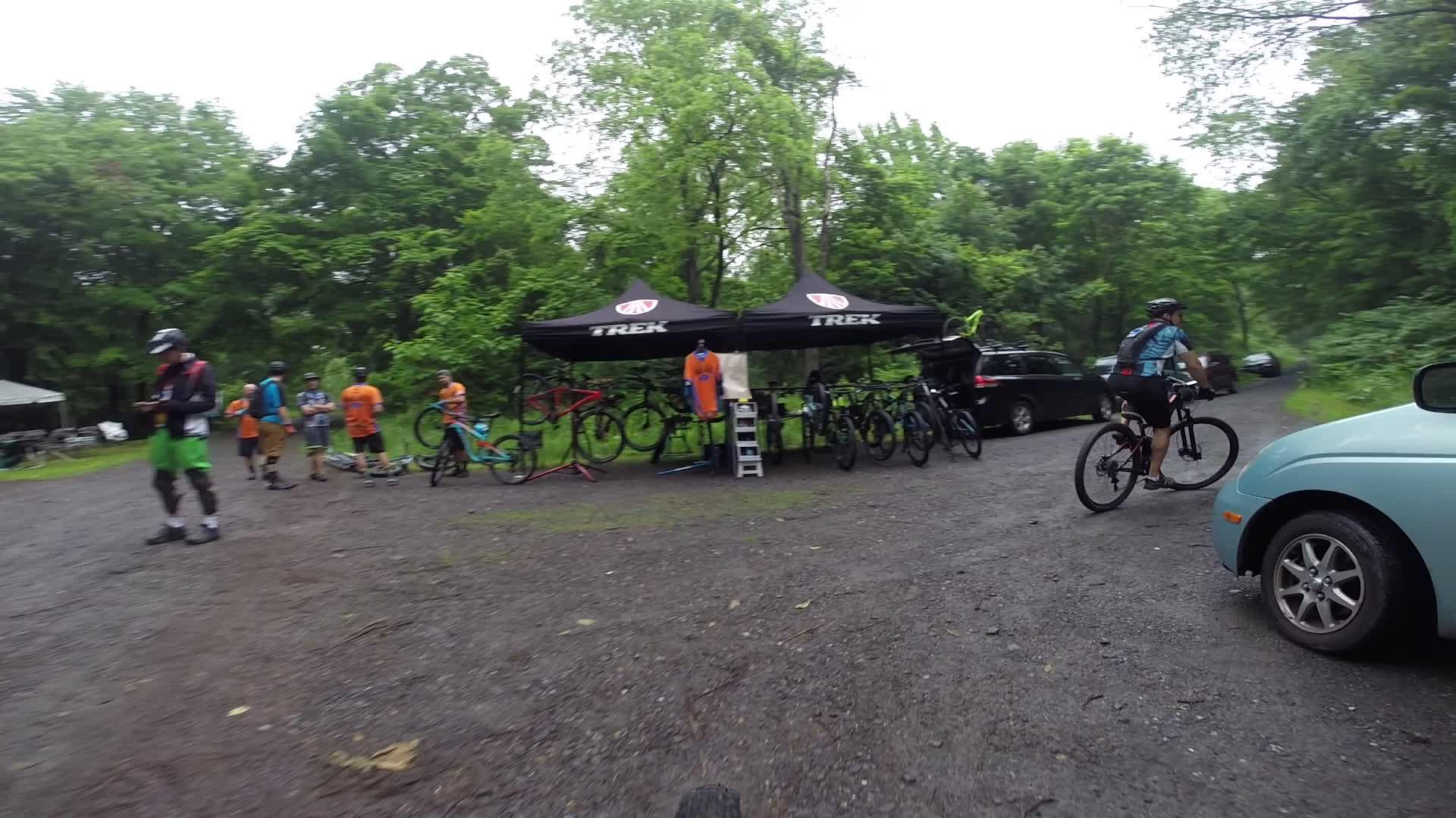 A mountain biking event at a wooded location with participants in bright orange shirts and cycling gear gathered near a TREK tent displaying bicycles. A cyclist in motion is departing the area, while several vehicles are parked along a gravel path. The scene is lush with greenery, indicative of a natural outdoor setting. Stewart State Forest mountain bike trail.