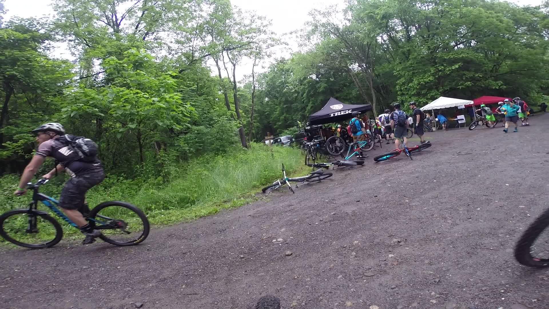 A mountain biker in motion on a gravel path, with lush greenery surrounding the scene. In the background, a group of people gather around tents and bicycles at a cycling event, under overcast skies. Some bikes are laid on the ground, while others are being inspected or ridden. Stewart State Forest mountain bike trail.
