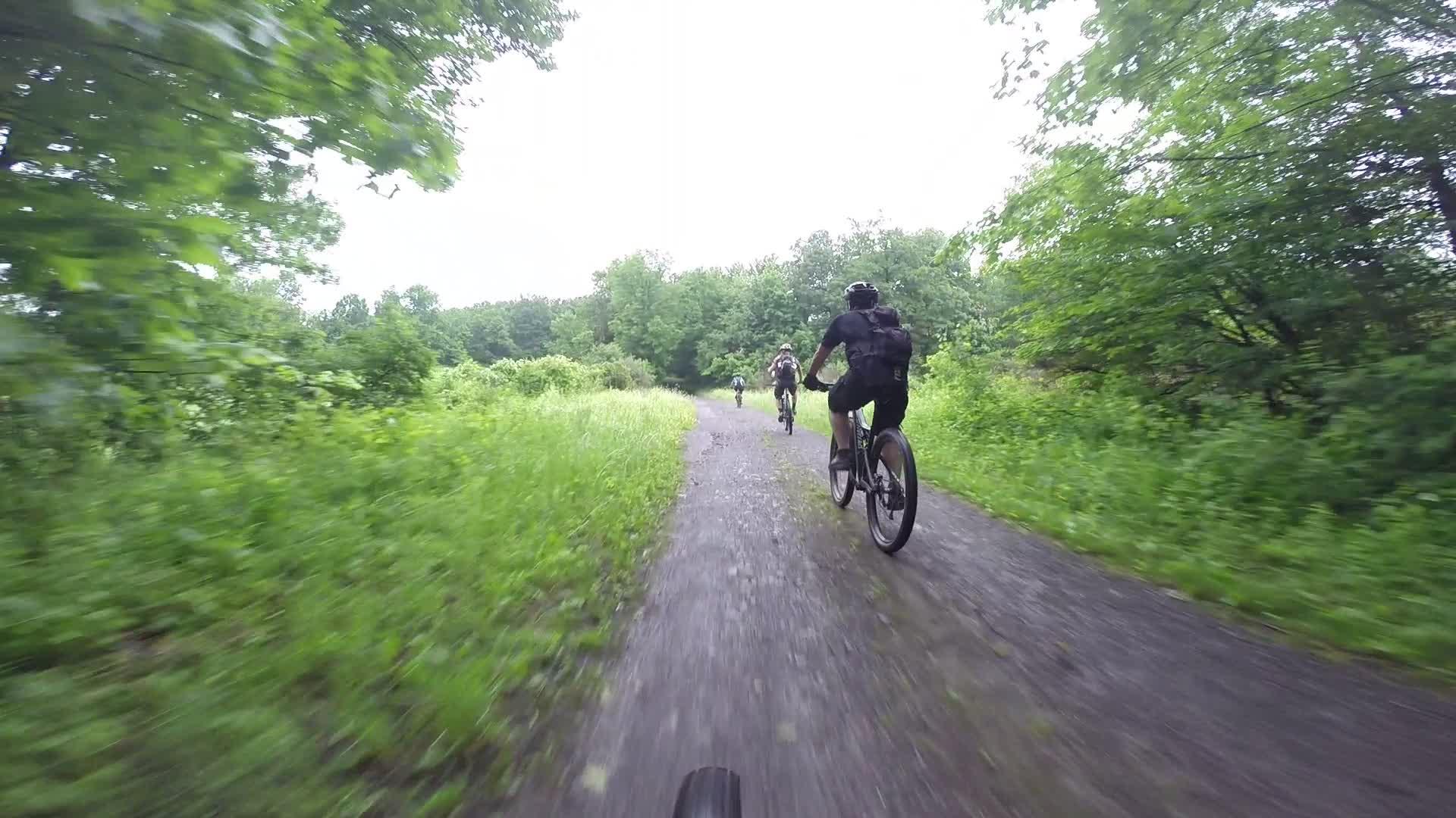 A view from the perspective of a cyclist riding on a gravel path, with two other cyclists in the distance. The path is surrounded by lush greenery and trees, indicating a forested area. The scene is overcast, suggesting a cloudy day. Stewart State Forest mountain bike trail.