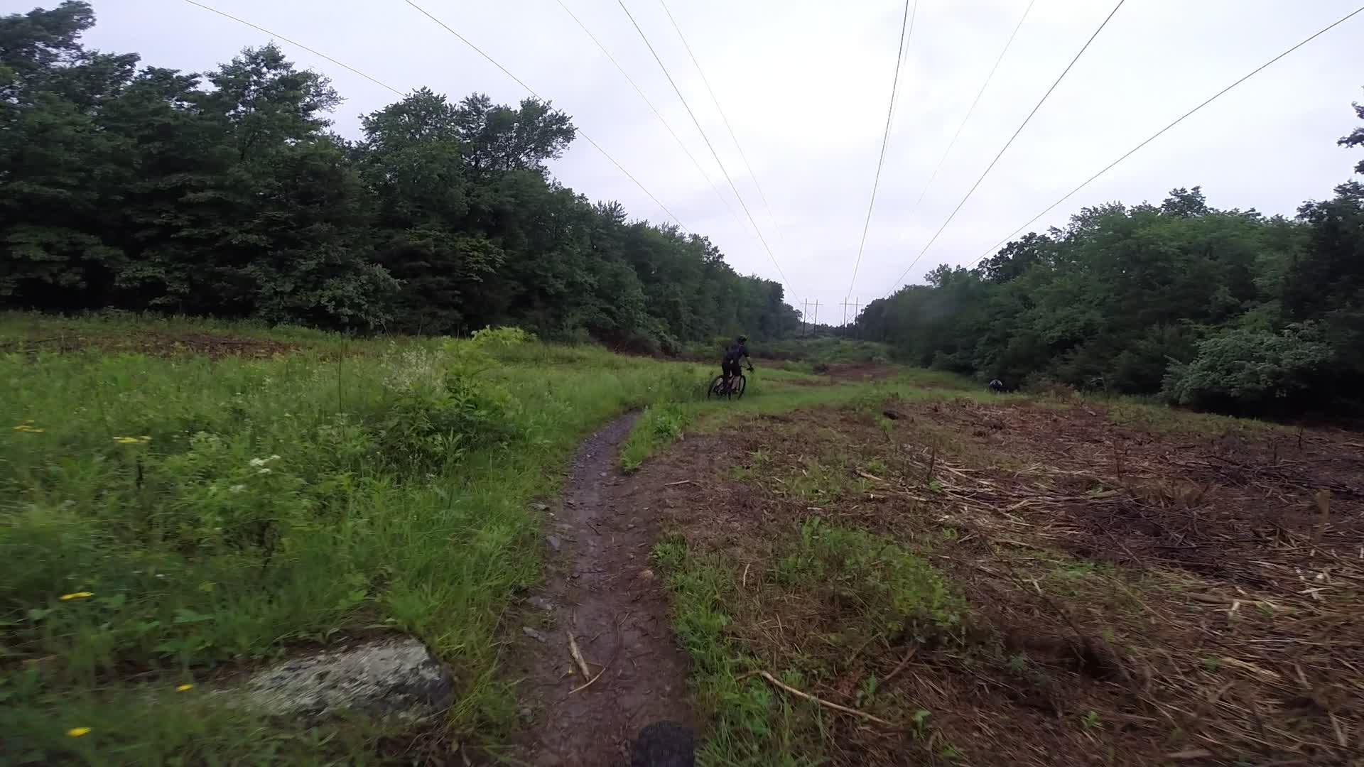 A cyclist riding along a muddy trail bordered by lush green grass and trees, with overhead power lines visible in the background. The scene is set on an overcast day, showcasing a natural environment that has been recently cleared in some areas. Stewart State Forest mountain bike trail.