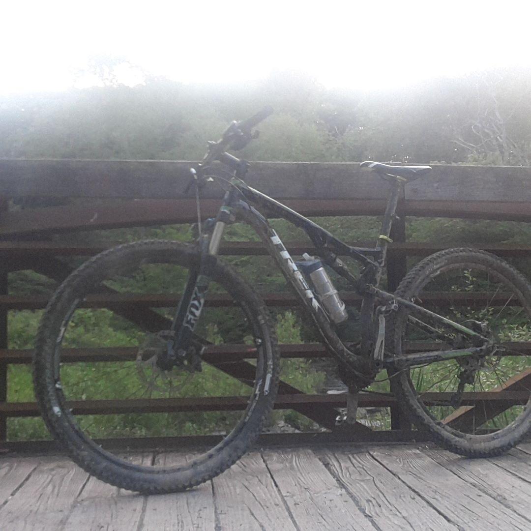 Trek Superfly FS 8: A mountain bike resting on a wooden bridge with a lush, green background. The bike has a black frame and muddy tires, indicating it has been used for off-road riding. A water bottle is attached to the frame.