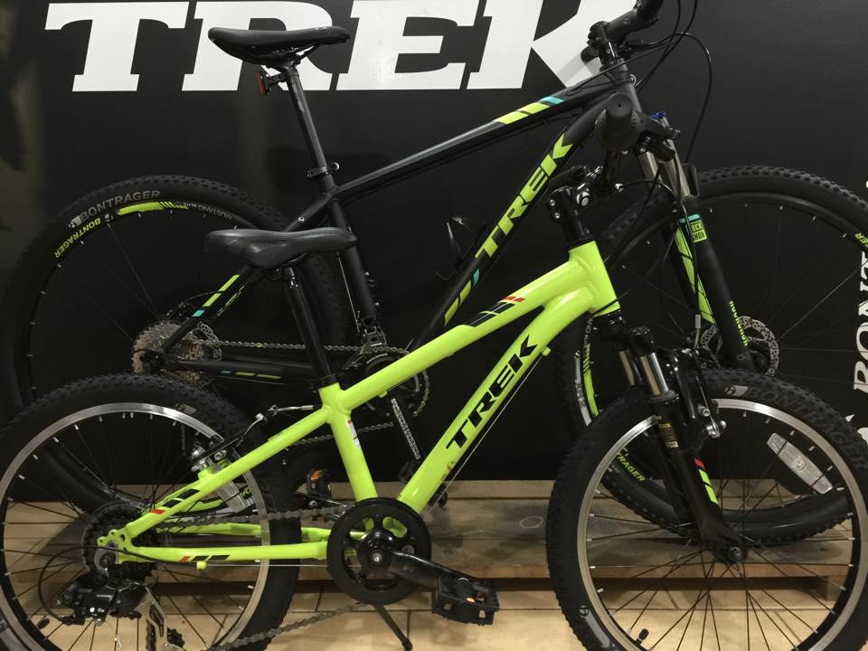 A bright green Trek bicycle is positioned in front of a black Trek bicycle, both showcasing distinct frames and tires. The background features a Trek logo prominently displayed. The image captures the bicycles from a side angle, highlighting their designs and colors.
