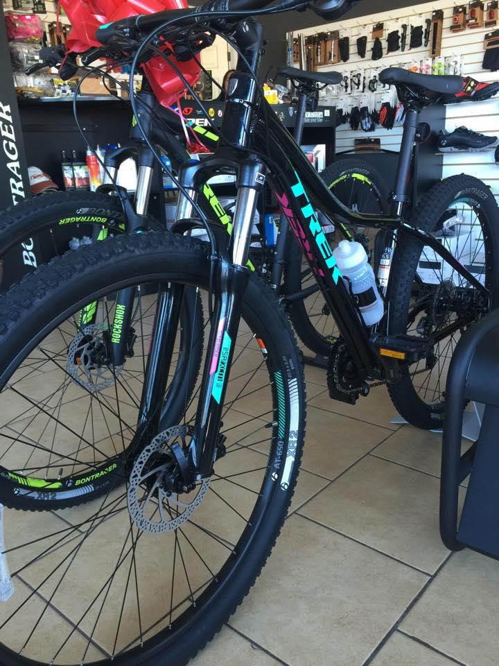 Alt text: A close-up view of two black mountain bicycles displayed in a bike shop. One bike is prominently in the foreground, showcasing its front wheel, fork, and handlebars, with colorful graphics and a water bottle attached. The shop interior features various bike accessories and parts in the background.