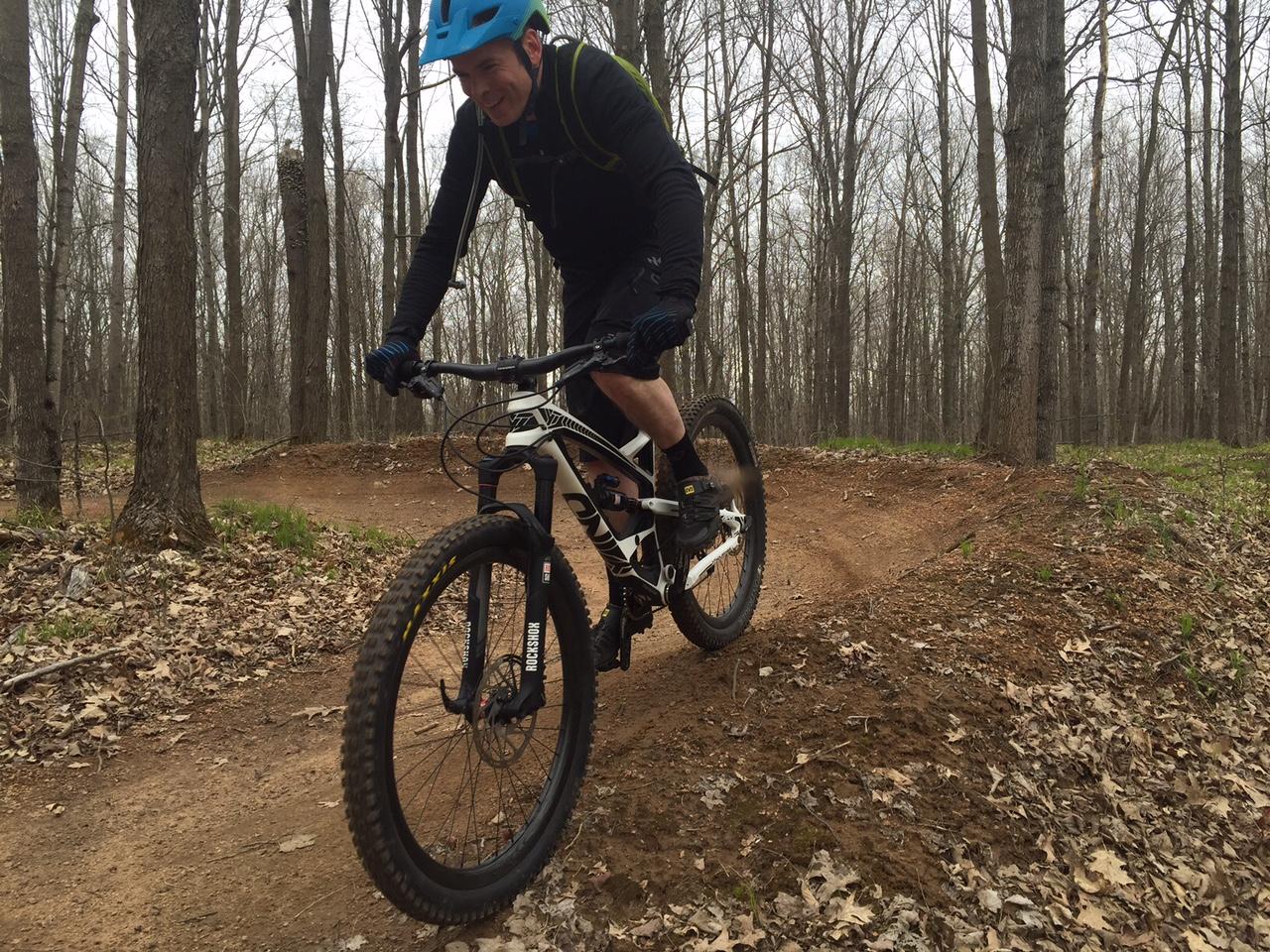 YT Industries Capra Carbon: A mountain biker navigating a dirt trail through a wooded area, wearing a blue helmet and cycling gear, with trees and fallen leaves in the background.