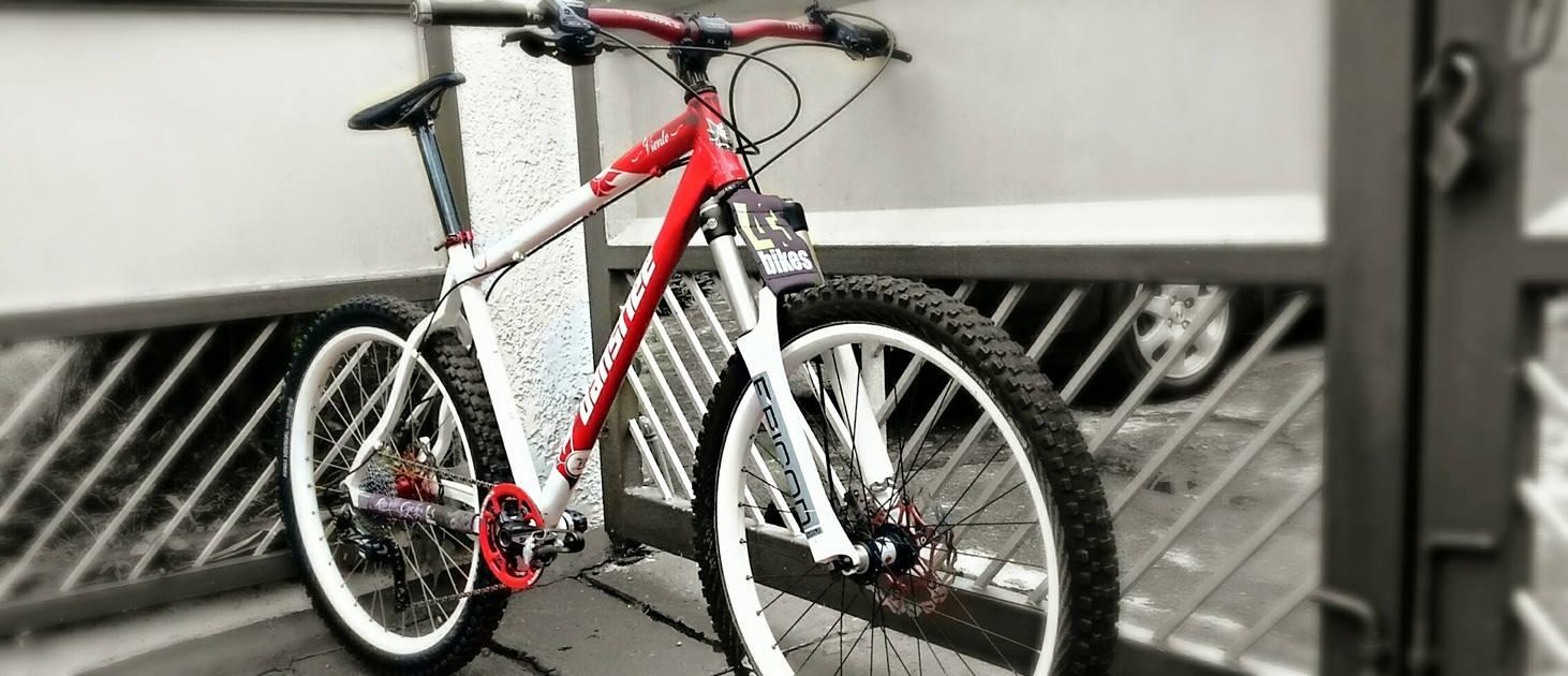 Banshee Viento: A mountain bike with a red and white frame is leaning against a gate. The bike features thick tires, a front suspension fork, and a visible gear mechanism. The background shows part of a gate and a blurred car. The image has a monochrome effect, emphasizing the bike's vibrant colors.