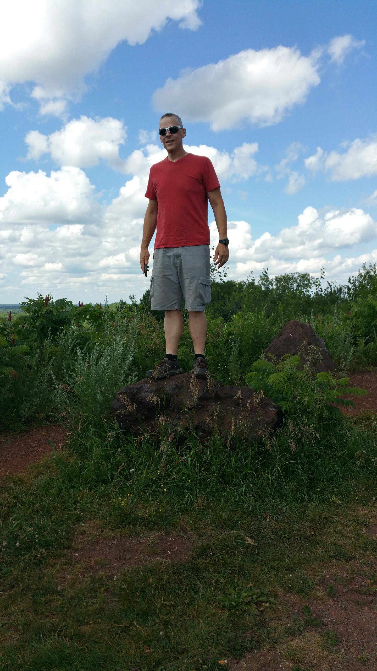 A person standing on a large rock in a grassy area, wearing a red t-shirt, shorts, and sunglasses. The background features a blue sky with fluffy clouds and greenery. Crusher/Miner's Mountain/Trammer/Ferrous wheel mountain bike trail.