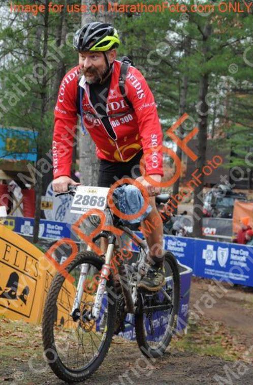 Jamis Dakar XC: A cyclist wearing a red jacket and black shorts pedals a mountain bike, smiling as he crosses a finish line at a cycling event. Surrounding him are banners and signs from various sponsors, with trees in the background. The cyclist's bib number is prominently displayed as he navigates the terrain.