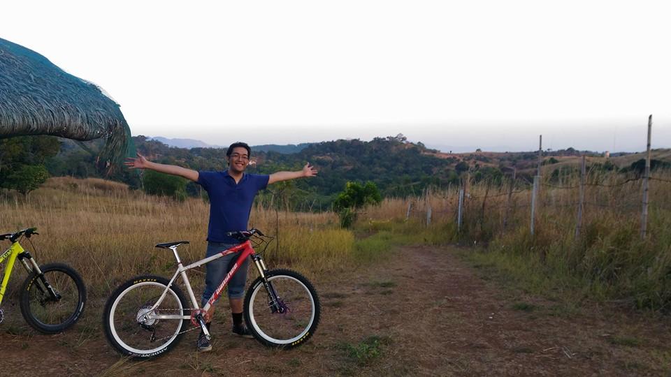 Banshee Viento: A person with outstretched arms is posing joyfully in a grassy landscape, surrounded by two mountain bikes. The background features rolling hills and a clear sky, suggesting a scenic outdoor location suitable for biking.