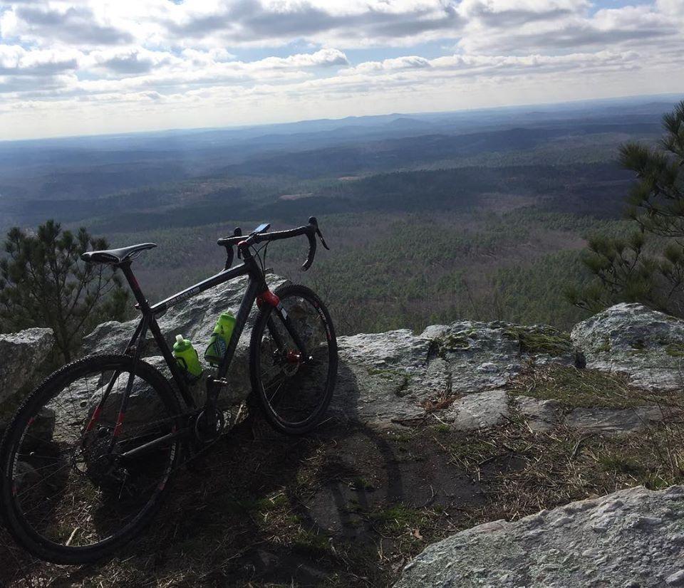 A black bicycle with red accents and two green water bottles is parked on a rocky outcrop overlooking a vast, forested landscape and distant mountains under a cloudy sky. Sylaward mountain bike trail.