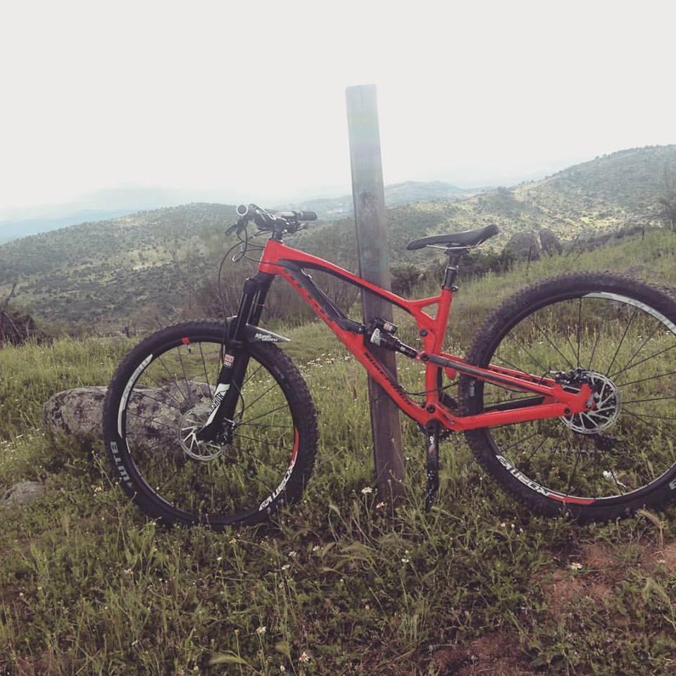 Nukeproof Mega: A red mountain bike resting against a wooden post on a grassy hillside, with rolling green hills and a cloudy sky in the background.