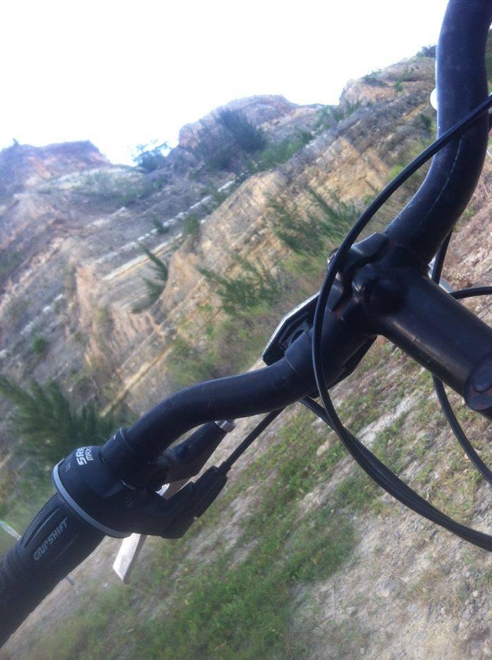 A close-up view of a bike handlebar with gear shift controls, set against a backdrop of a rugged landscape featuring steep, rocky slopes and sparse vegetation. Monte Realengo mountain bike trail.